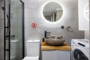 Uma casa de banho moderna com chuveiro em cabine de vidro, espelho redondo, lavatório com bacia de pedra, sanita e máquina de lavar roupa.
