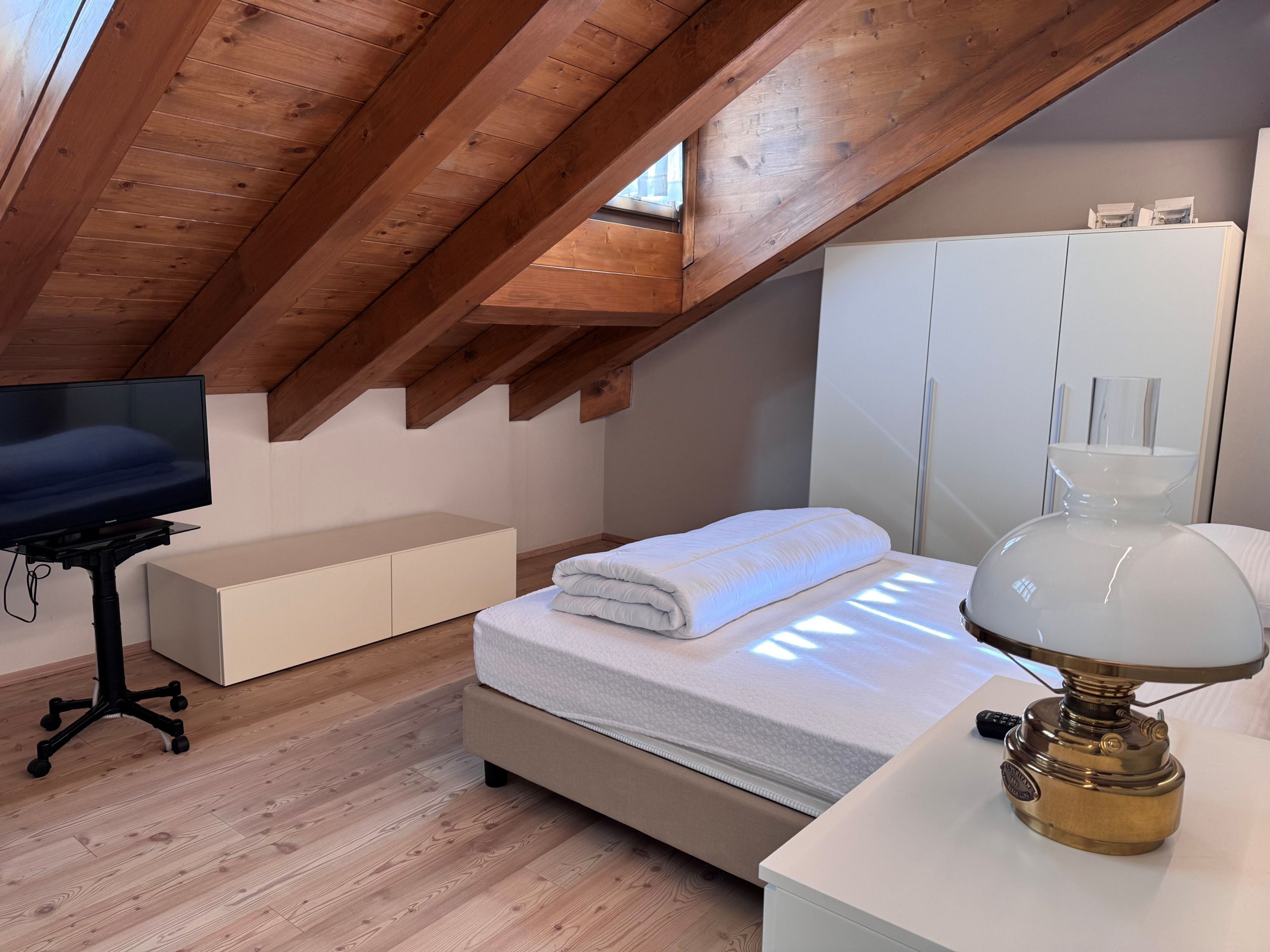Un dormitorio con techo inclinado, una cama con edredón blanco, una lámpara de noche, un televisor sobre un mueble bajo y un armario.
