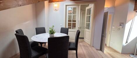 Un comedor con una mesa redonda y seis sillas, una planta en maceta, y vista a una cocina y comedor a través de un umbral abierto.