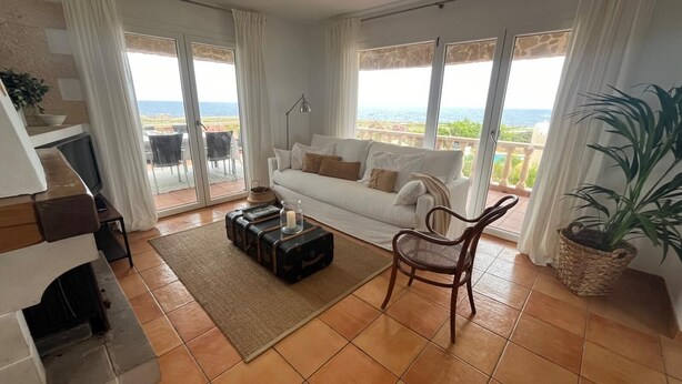 A living room with a sofa, a chair, a fireplace, a table with a lamp, and a view of the sea through large windows. - Villa Miramar. Spectacular views. (Es Mercadal)