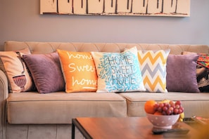 A beige sofa with four decorative pillows, a bowl of fruit, and a remote control on a wooden table.