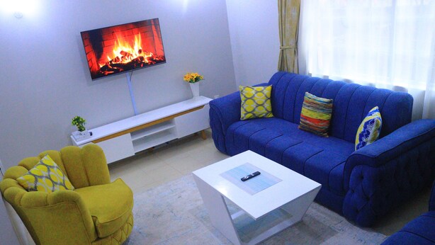 A living room with a blue sofa, a yellow armchair, a white coffee table, a flat-screen TV, and a small white shelf with a plant.