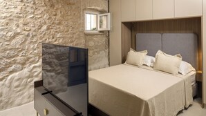 A bedroom with a stone wall, a bed with a grey headboard, and a TV on a dark dresser. - Villa Maja Aurelia in the Centre- Romantic Studio Apartment (Hvar)