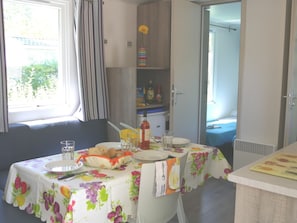 Uma área de refeições com uma mesa posta para dois, uma janela com cortinas e uma cozinha compacta ao fundo.