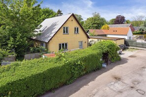 A yellow house with a sloped roof, surrounded by greenery and a paved driveway.