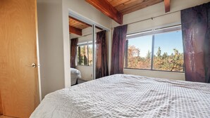 Une chambre avec un grand lit, des poutres apparentes en bois au plafond, une fenêtre avec des rideaux et une vue sur les arbres à l’extérieur.