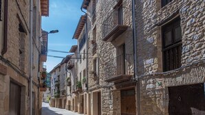 Una calle estrecha flanqueada por edificios de piedra, con balcones adornados con plantas en macetas, y un cielo despejado arriba.