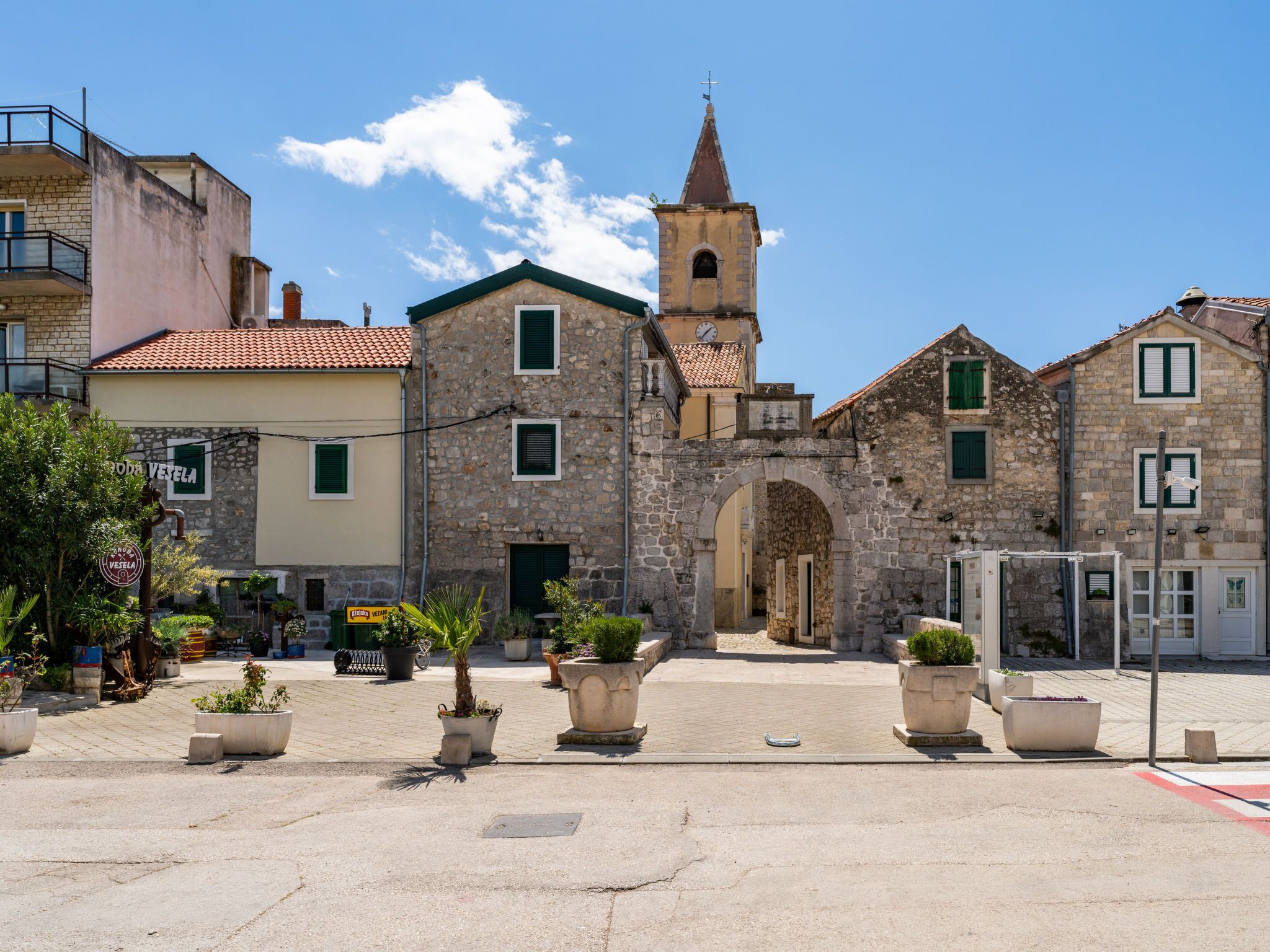 A historic stone building with a clock tower, arched entrance, and a paved square with potted plants.