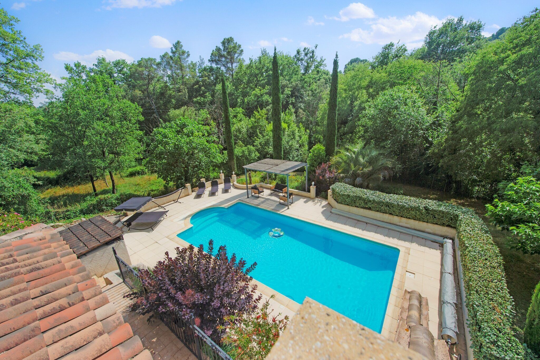 A rectangular swimming pool with a sunshade, surrounded by a landscaped garden with hedges and trees.