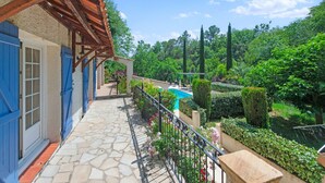A stone-paved balcony with a wooden pergola, a swimming pool, and lush greenery.
