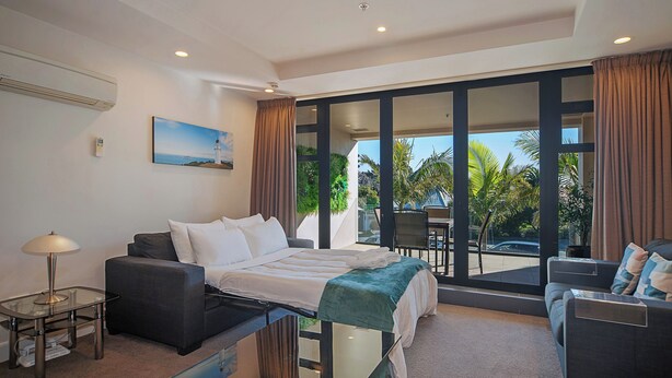 A modern hotel room with a bed, a sofa, a glass table, and a view of palm trees and a car through large windows.