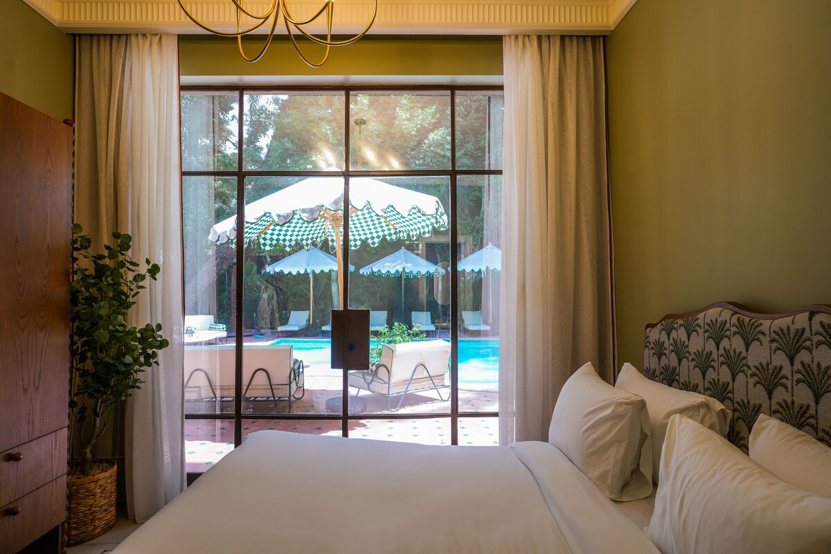 A hotel room with a large window, a bed with white linens, a wooden cabinet, and a potted plant.
