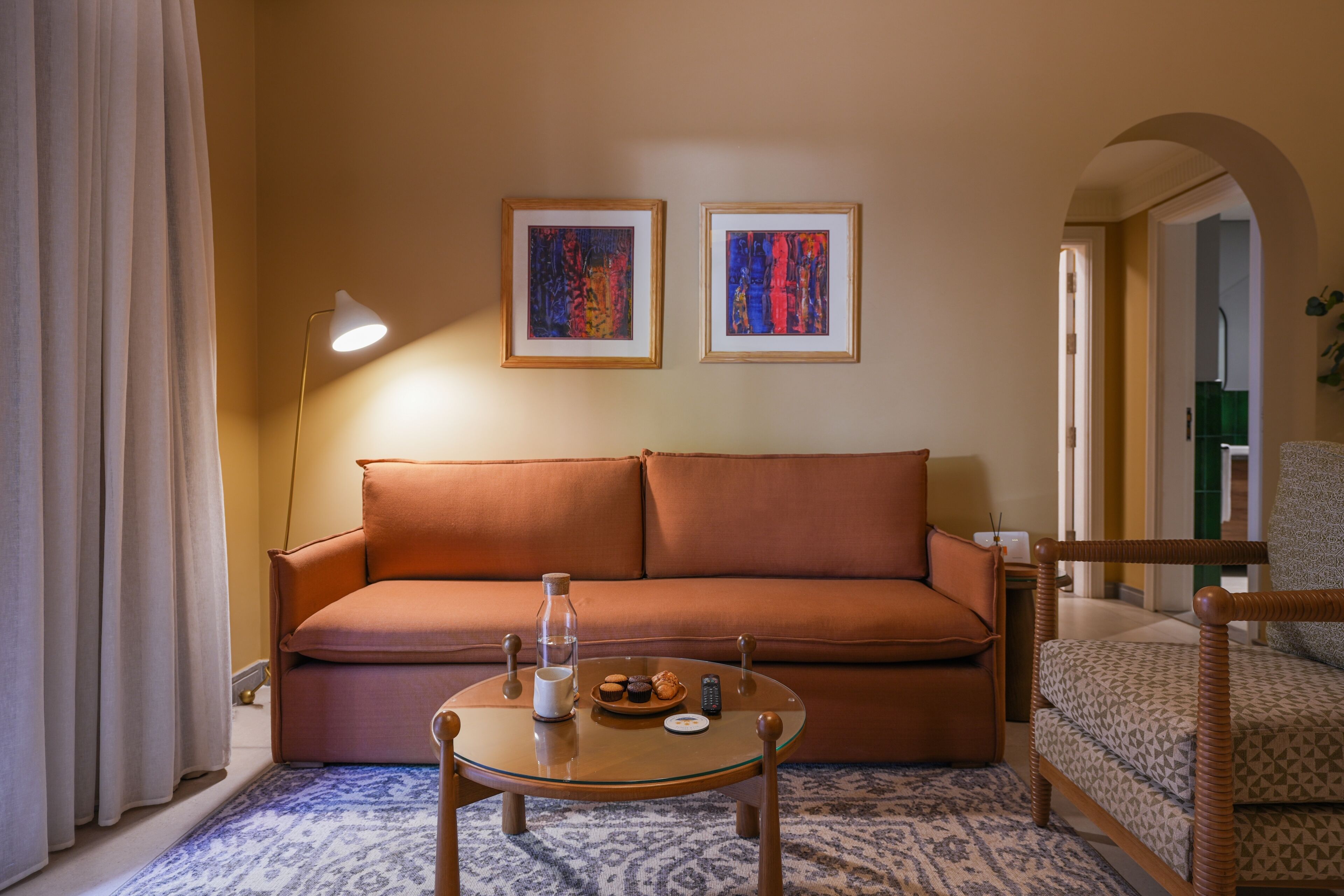 A room with a sofa, a small round table with a pitcher, a remote, and a plate of cookies. There are two framed artworks on the wall.