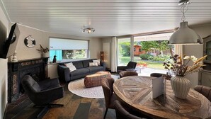 Ein modernes Wohnzimmer mit Kamin, einer Couch, einem Esstisch und einem großen Fenster mit Blick ins Freie.