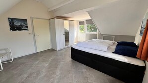 Ein Schlafzimmer mit einem großen Bett, einem Kleiderschrank, einer Tür zu einem anderen Raum und einem Fenster mit Blick ins Grüne.