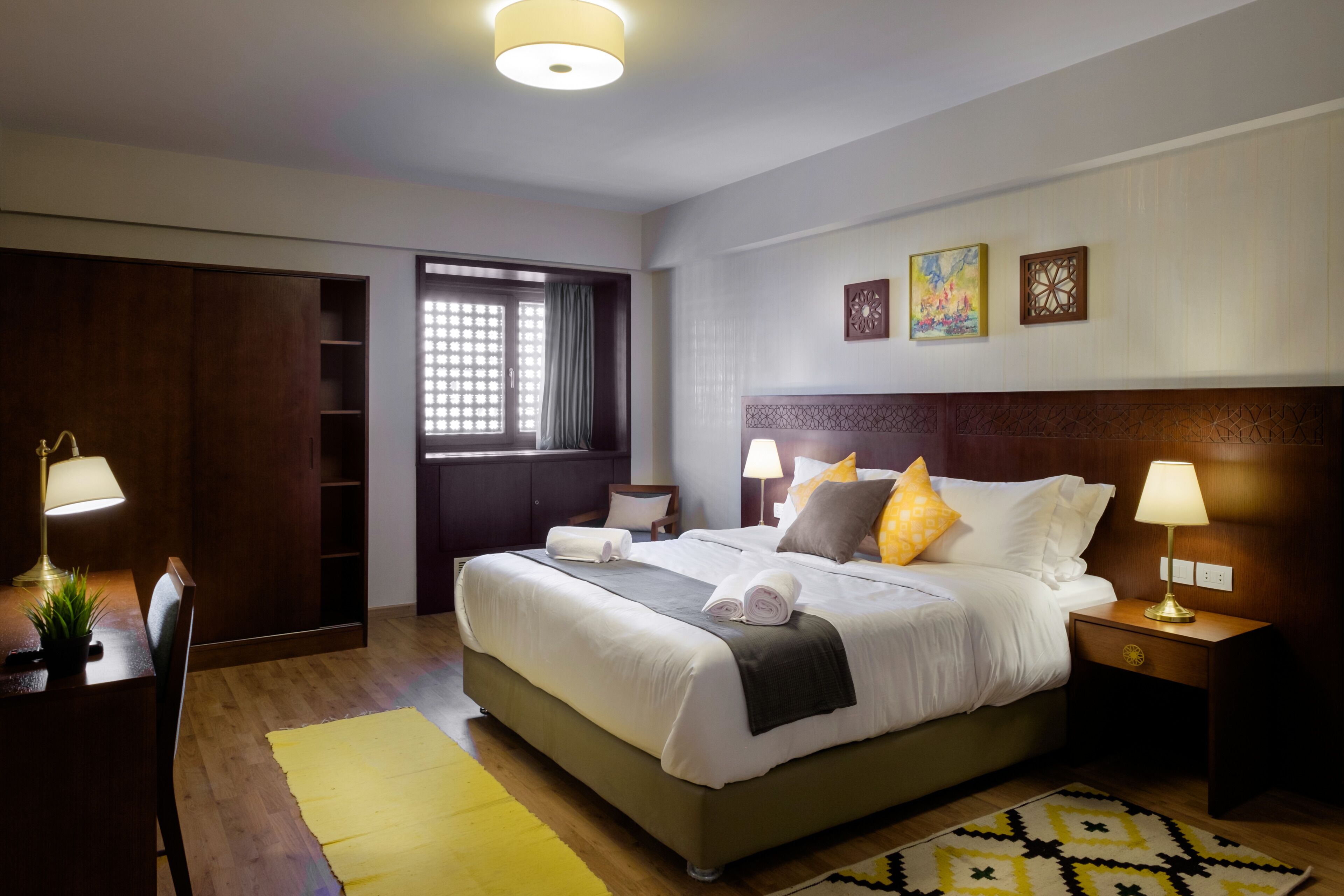 Une chambre d’hôtel avec un grand lit, des lampes de chevet, un bureau avec une lampe, une chaise, une fenêtre avec des rideaux et des œuvres d’art accrochées au mur.