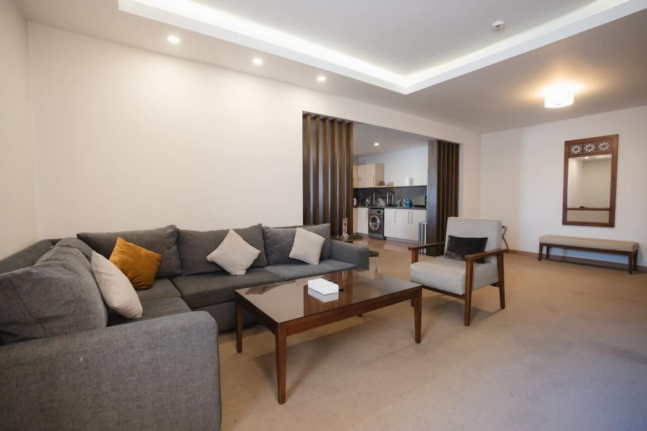 Una sala de estar moderna con un sofá gris, una mesa de centro de madera y una silla. Se aprecia una cocina al fondo con armarios y un lavamanos.