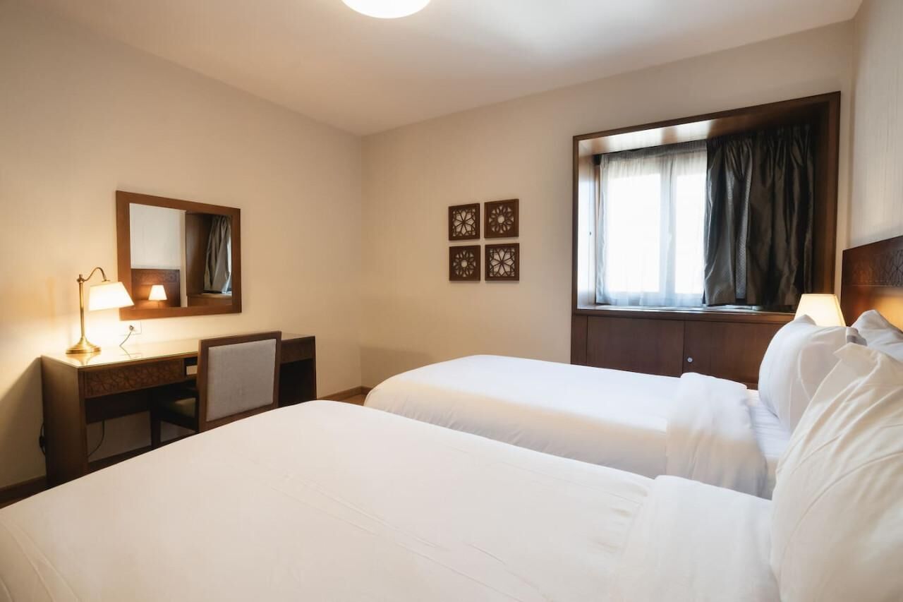 Een hotelkamer met twee bedden, een bureau met lamp, een spiegel en een raam met gordijnen.