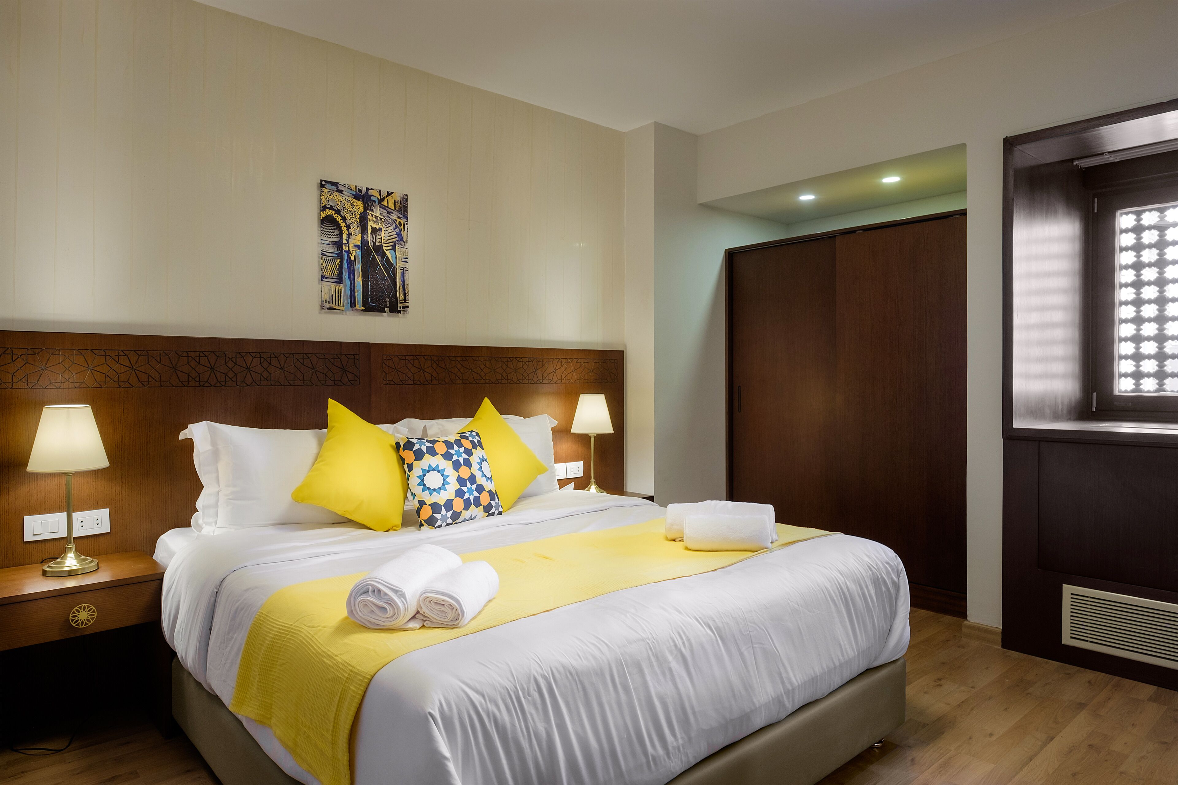 Habitación de hotel con una cama grande, dos lámparas de noche, cabecero de madera, un elemento decorativo en la pared y una ventana con persianas.