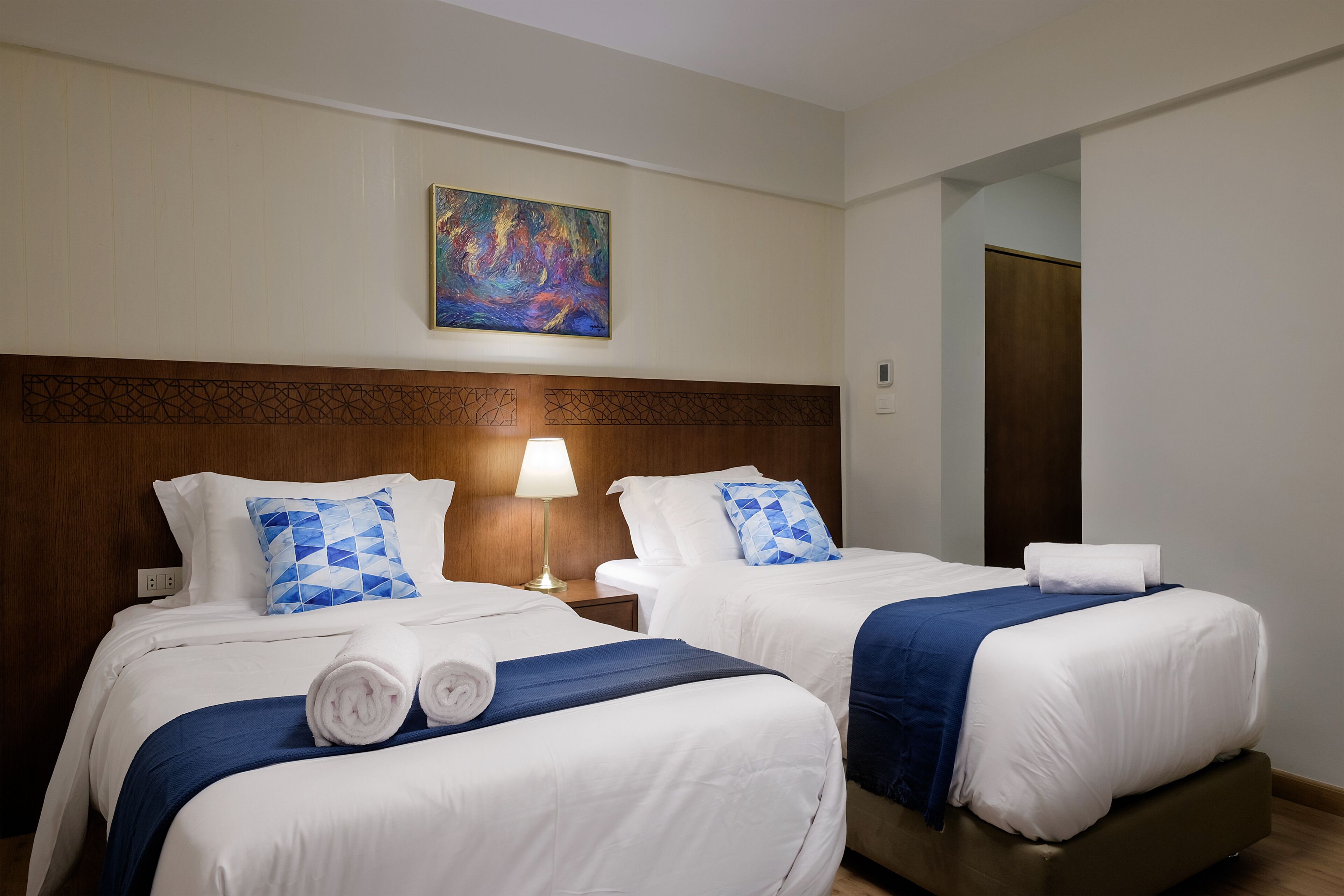 A hotel room with two beds, a wooden headboard, a painting on the wall, and a nightstand with a lamp.