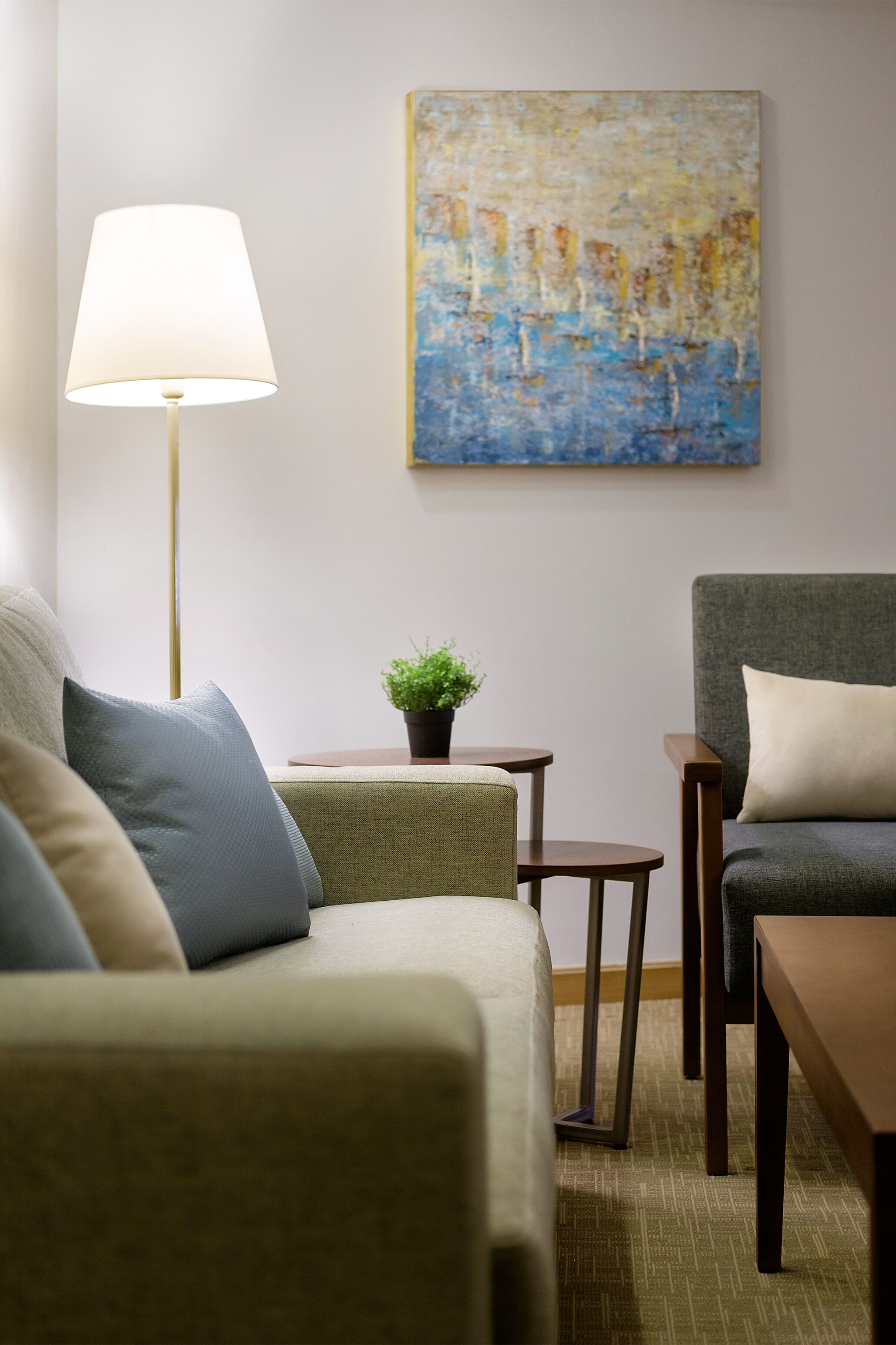 A room with a sofa, armchair, small table, lamp, and a painting on the wall.