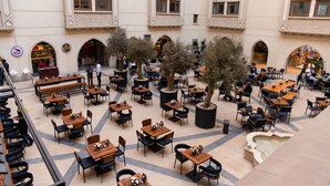 Ein Straßencafé mit mehreren Tischen und Stühlen, umgeben von Bäumen und architektonischen Elementen.