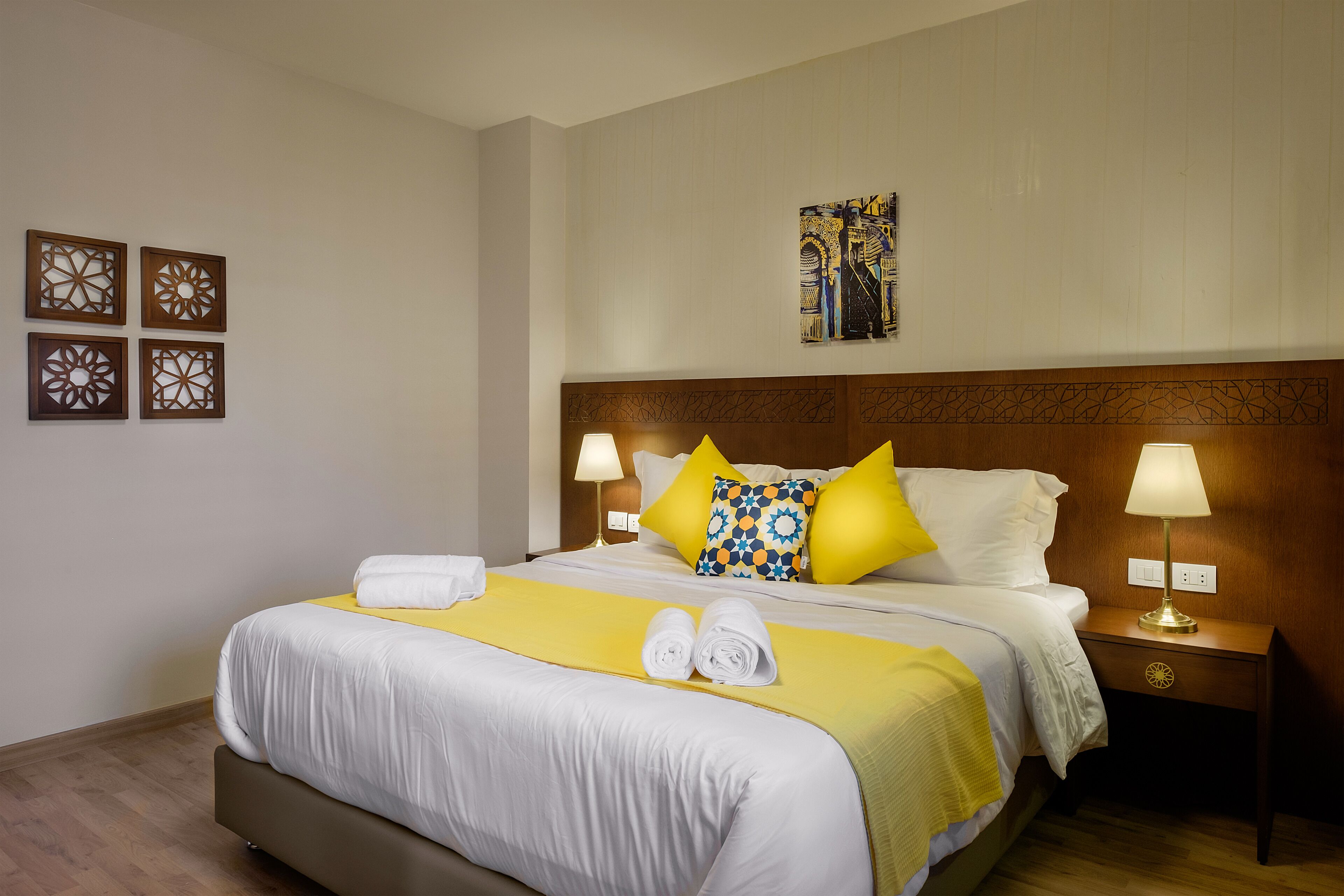 A neatly made bed with white linens, a yellow throw blanket, and two rolled towels. There are two bedside lamps, a wooden headboard, and a wall-mounted artwork above the bed.