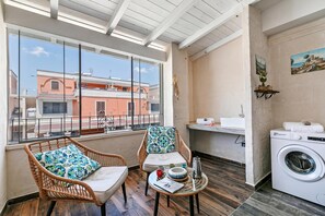 Un balcon avec des fauteuils en osier, une petite table avec un livre, un lave-linge et une vue sur les immeubles.