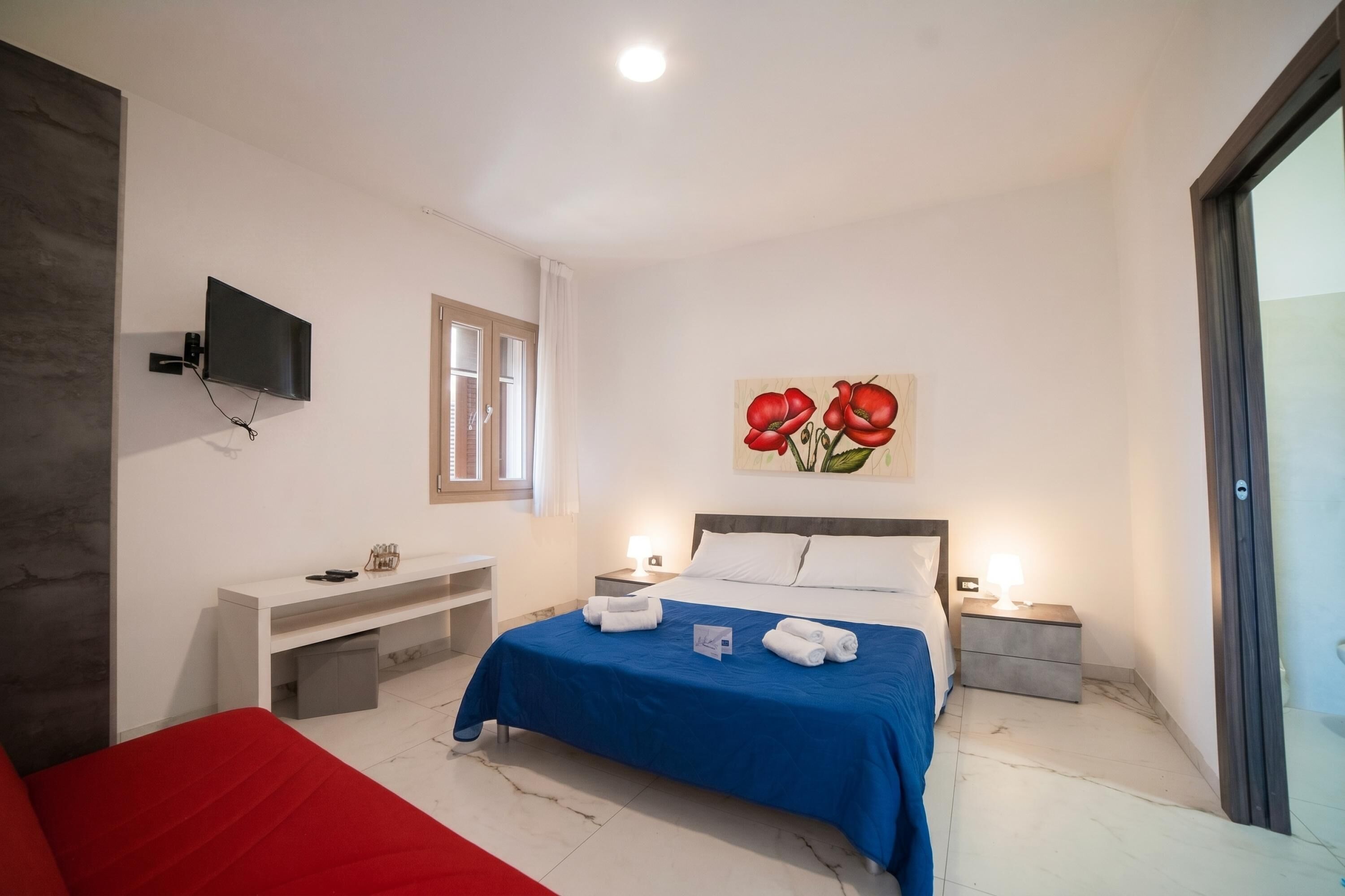 Quarto de hotel com uma cama, televisão fixada na parede, escrivaninha, mesa de cabeceira e um quadro com flores vermelhas.
