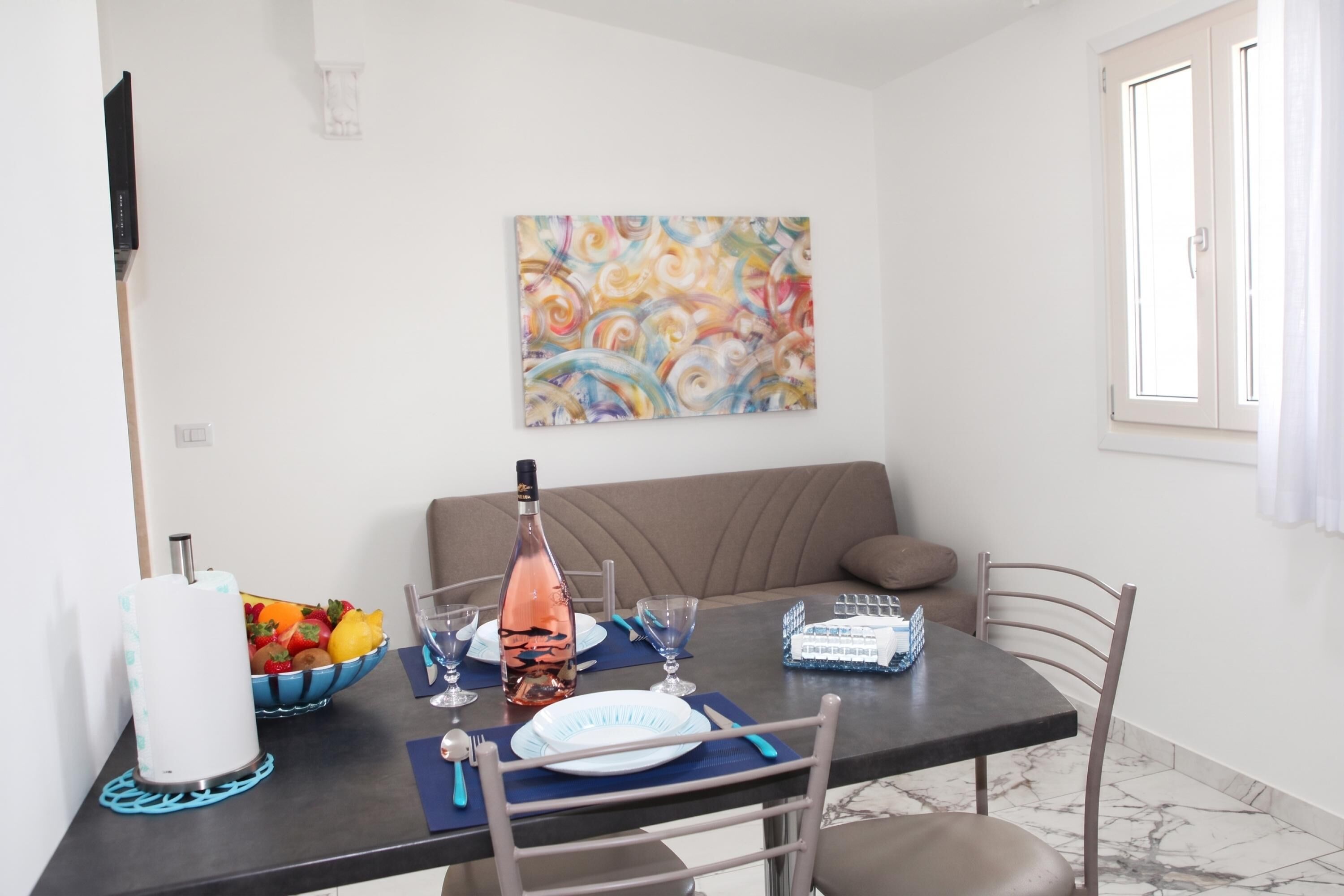 Un comedor con una mesa dispuesta para dos, una botella de vino, un frutero y un cuadro en la pared.