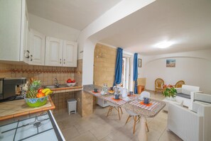 Uma cozinha compacta com armários brancos, um espaço de refeições com toalha quadriculada e uma área de estar com móveis de vime.