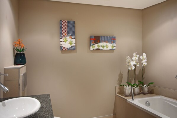 A bathroom with a sink, mirror, and two wall-mounted paintings. There are orchids in vases on the counter and a bathtub. - Cape Eazi Stayz The Rockwell (Cape Town)