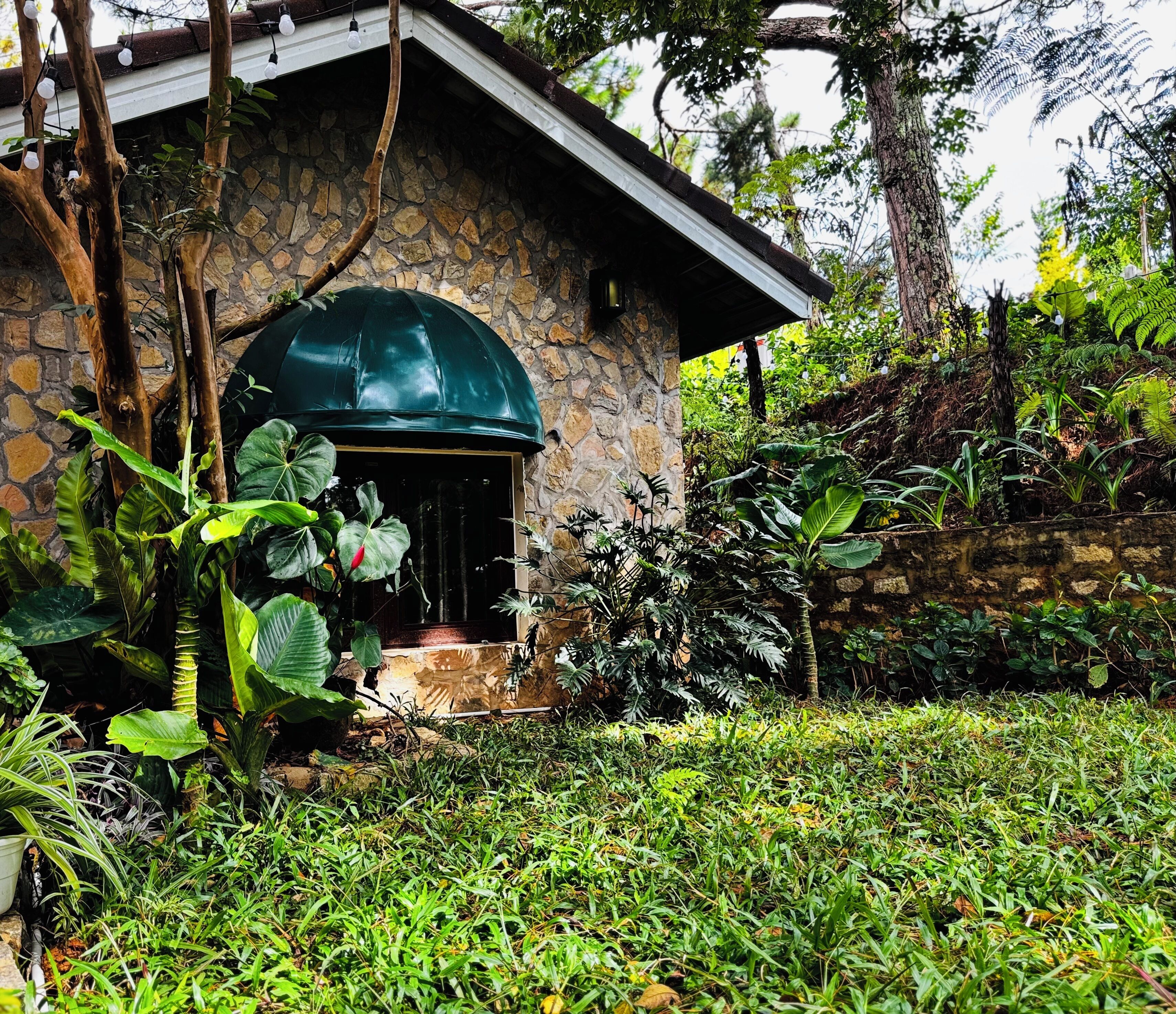 A stone building with a green dome roof, surrounded by lush greenery and a stone wall.