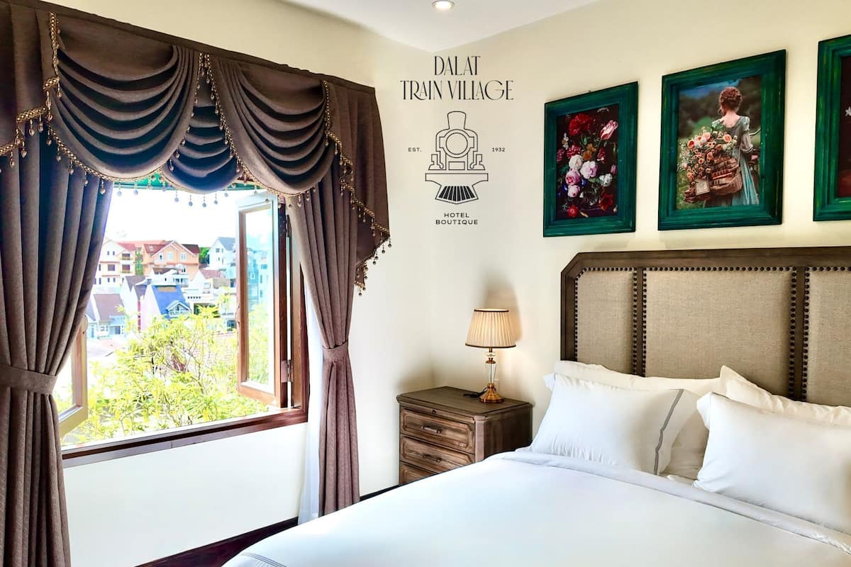 Habitación de hotel con una cama grande, una mesita de noche con lámpara, vista al pueblo por la ventana y una pared con el nombre del hotel y su año de fundación.
