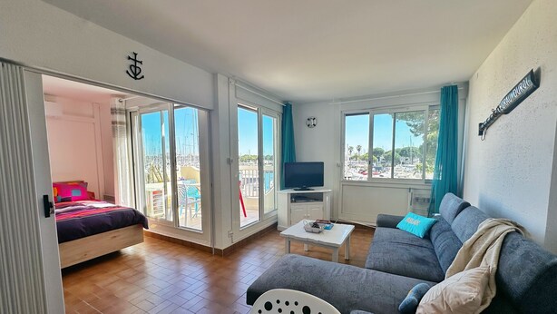 A room with a bed, a sofa, a TV, and a balcony with a view of boats. - Sun, peace & comfort in Port Camargue (Le Grau-du-Roi)