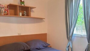 Een enkel bed met houten hoofdbord, een grijze spreideken en een opgevouwen deken. Boven het bed bevindt zich een plankje met enkele spullen, waaronder een beeldje.