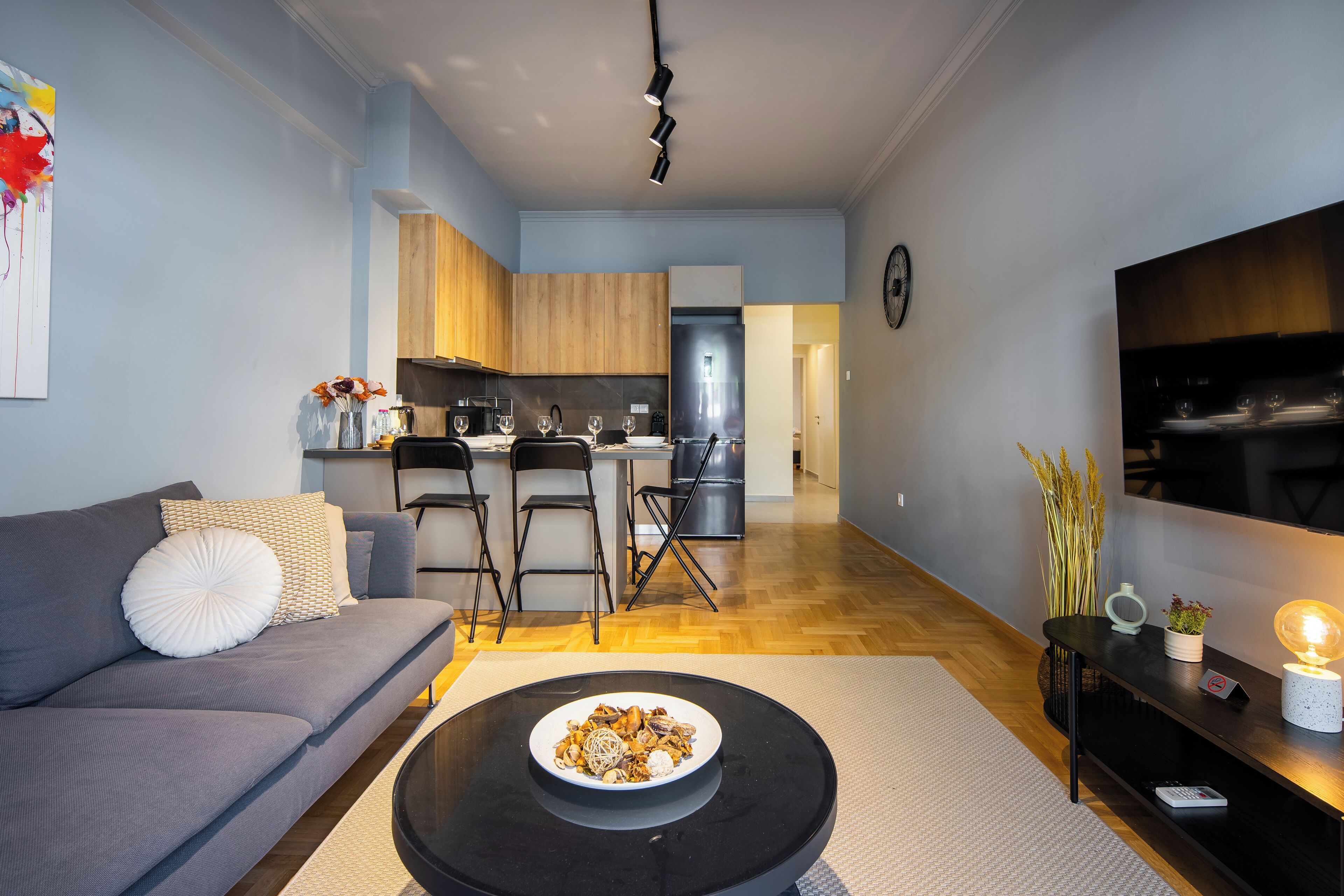 Un salon moderne avec un canapé gris, une table basse ronde sur laquelle se trouve un plateau, un meuble TV noir avec une lampe, et une cuisine en arrière-plan.