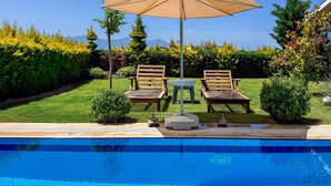 A poolside area with two wooden lounge chairs, a table, and a large umbrella. - Villa Limon (Kusadasi)