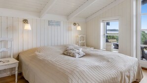 Une chambre à coucher avec un grand lit, deux lampes fixées au mur, une petite table et une vue sur l’extérieur par une fenêtre.