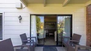 Eine Terrassenfläche mit zwei Korbstühlen und einem kleinen Tisch, eine Glastür, die zu einem Innenbereich führt, und eine Backsteinmauer an einer Seite.