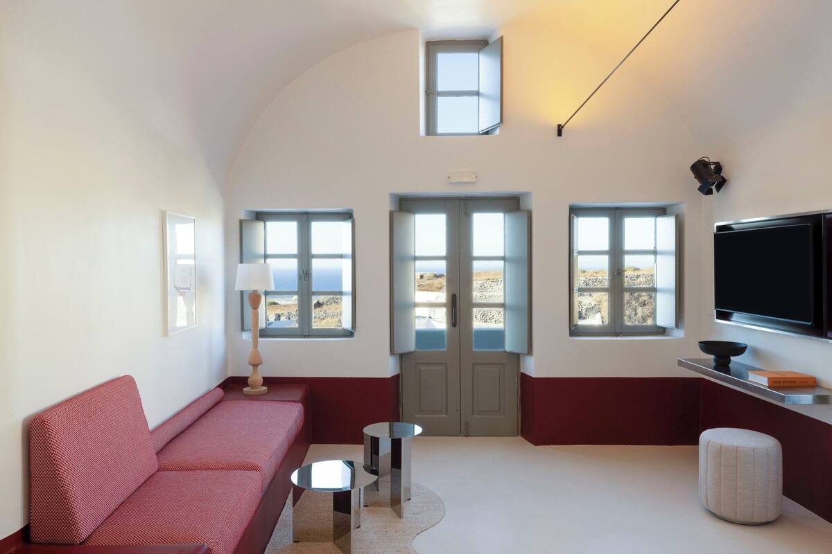 Een moderne woonkamer met een rode bank, een kleine ronde tafel, een televisie en een deur naar een balkon met uitzicht op zee.