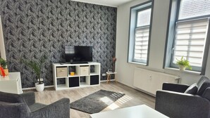 A living room with a patterned wallpaper, a flat-screen TV on a white entertainment unit, grey sofas, a white coffee table, and a potted plant by the window.