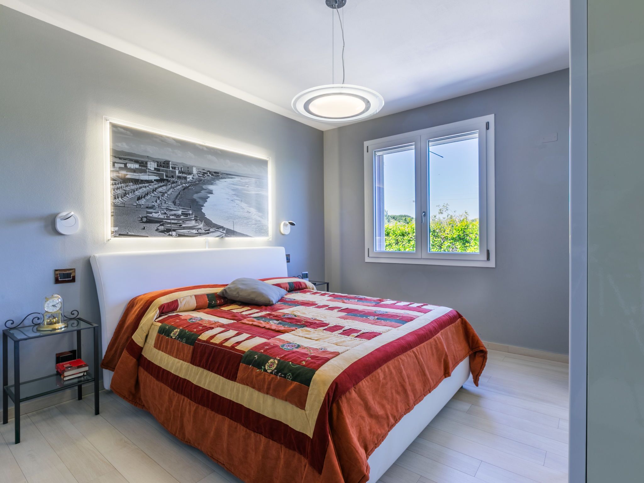 A bedroom with a large bed, a nightstand, a wall-mounted light, a window with a view of greenery, and a framed picture on the wall.