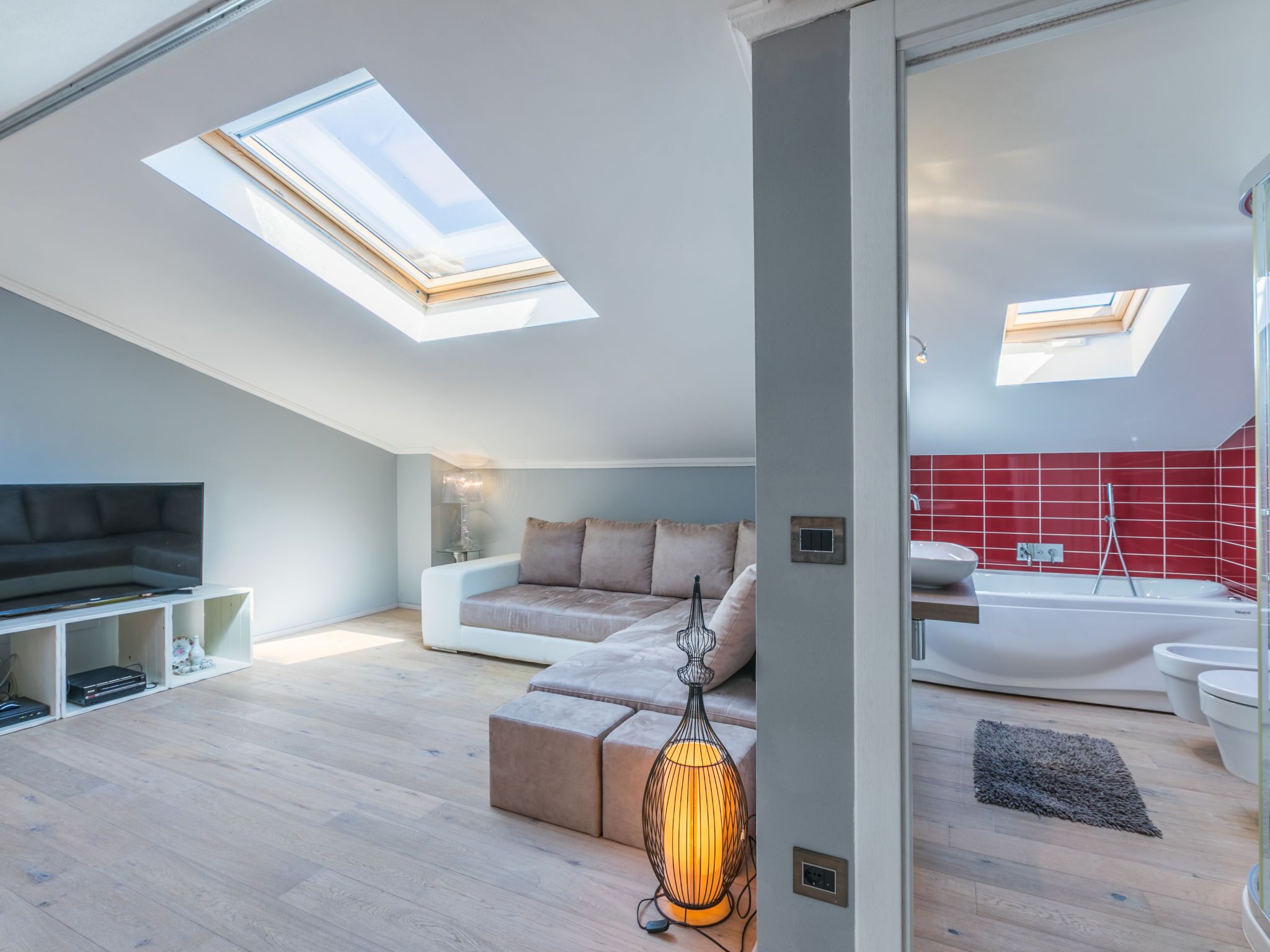 Un appartement moderne de style « loft », doté de fenêtres de toit, d’un espace de vie avec un canapé et un téléviseur, et d’une salle de bain avec une baignoire et des carreaux rouges.