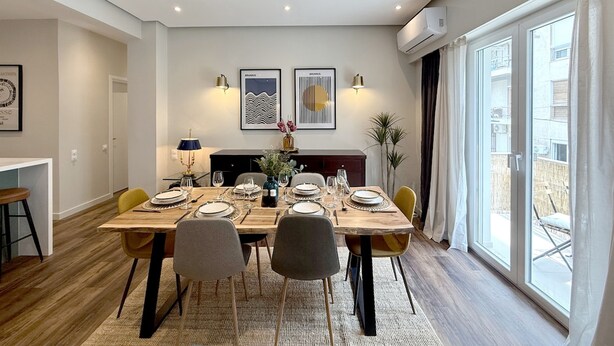 A dining area with a wooden table set for four, surrounded by chairs, with artwork on the wall and a view of the outdoors through large windows. - Siga-siga Luxury Acropolis Apt (Athens)