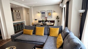 A modern living room with a grey sectional sofa, a black coffee table, and a dining area in the background. - Siga-siga Luxury Acropolis Apt (Athens)