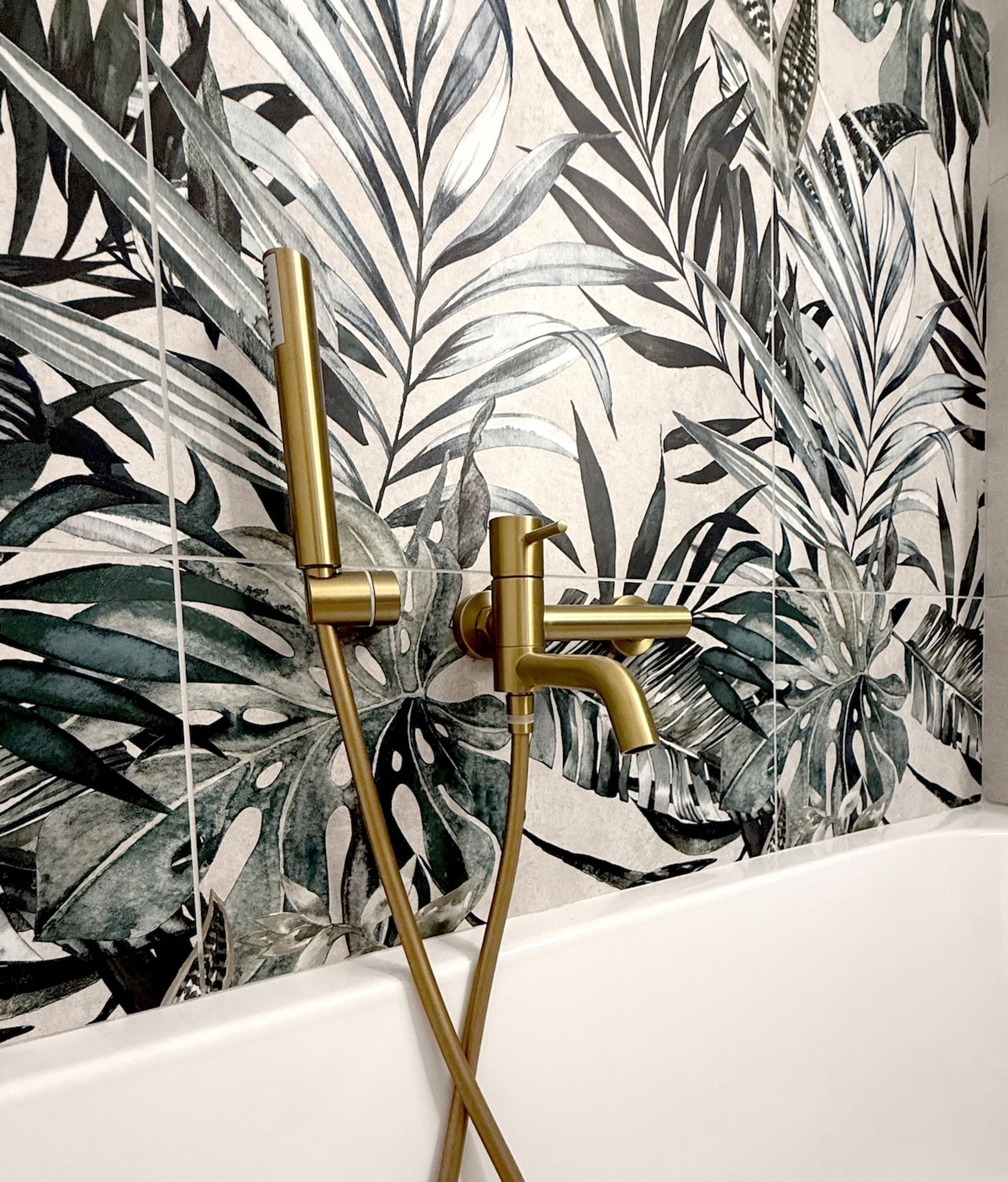 Une salle de bain avec une baignoire, des accessoires de douche de couleur dorée et un mur orné d’un motif de feuilles tropicales.