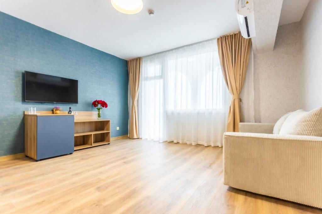 A modern living room with a flat-screen TV, a wooden cabinet, a beige sofa, and a large window with sheer curtains.
