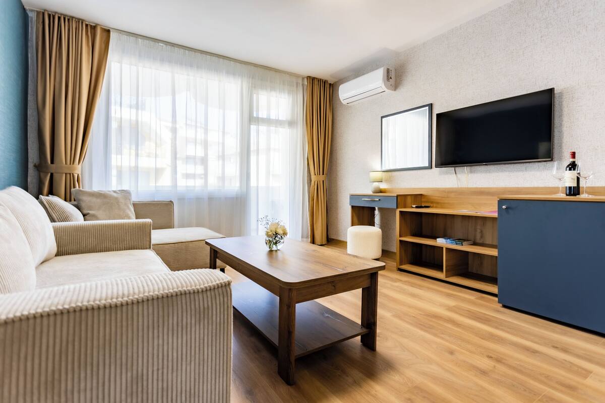 A modern hotel room with a sofa, a wooden coffee table, a TV, and a minibar.