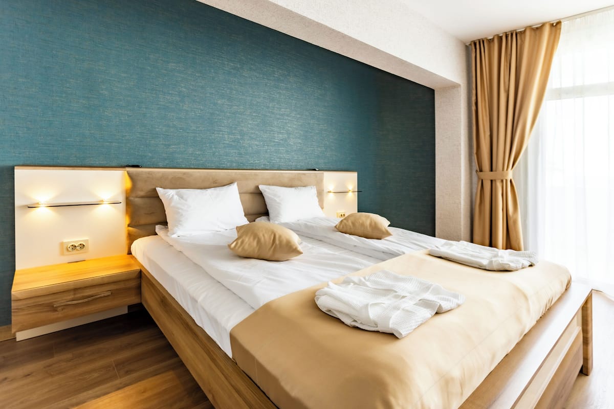 A hotel room with a large bed, wooden headboard, bedside tables with lamps, and a view of the outdoors through a window with curtains.